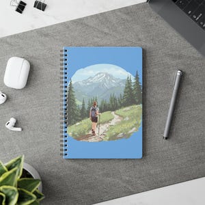 May include: Spiral-bound notebook with a landscape illustration of a hiker on a mountain trail. The cover shows a sunny scene with green trees, a blue sky, and snow-capped mountains. The notebook is on a grey surface with other office supplies.