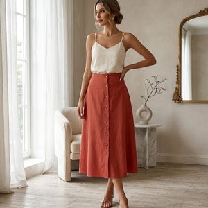May include: A woman wearing a rust-colored button-down midi skirt and a cream-colored camisole. The skirt has a high waist and a row of buttons down the front. The outfit is paired with strappy sandals.