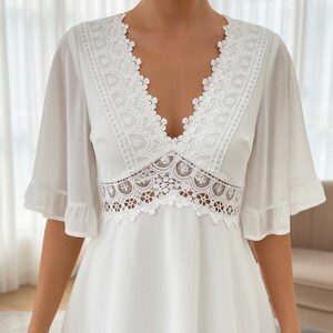 Puede incluir: Vestido blanco con escote en V y mangas cortas con volantes. El vestido presenta intrincados detalles de encaje blanco alrededor del escote y la cintura, creando un aspecto delicado y elegante. La tela parece ser un material ligero y fluido.