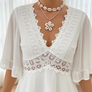 Puede incluir: Un vestido blanco con escote en V y detalles de encaje. El vestido presenta mangas cortas con bordes con volantes y un panel de encaje decorativo en la cintura. Un collar en capas con colgantes de concha a&ntilde;ade un toque de elegancia.