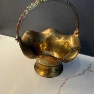 May include: A polished brass basket with a scalloped, wavy rim and a matching arched handle. The basket features a wide, flared opening that tapers down to a circular pedestal base, showcasing a metallic gold finish with subtle reflective surfaces.