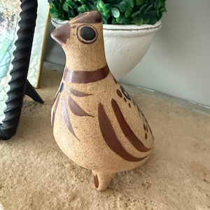 May include: A ceramic bird figurine in shades of tan and brown. The bird has a rounded body, a small tail, and a long neck with a detailed head. Brown stripes and accents decorate the body. The bird is a decorative item.