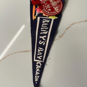 May include: A dark blue felt pennant featuring the white text "BARRY'S BAY CANADA" and a red circular patch at the top with the text "CAMP KAKWITHA" and a graphic illustration of a person. A small red ribbon is attached to the top corner.