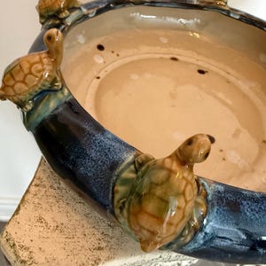 Puede incluir: Un cuenco de cerámica con un esmalte azul y beige, adornado con figuras de tortugas alrededor del borde. Las tortugas son marrones y verdes, añadiendo un toque caprichoso a la pieza decorativa. El cuenco es probablemente decorativo.