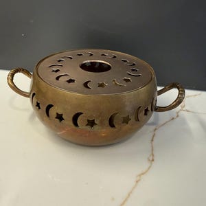 May include: A round brass warmer featuring two side handles and a removable lid. The body and lid are decorated with cut-out shapes of crescent moons and stars. The metal has a darkened, aged patina finish throughout the entire exterior surface.