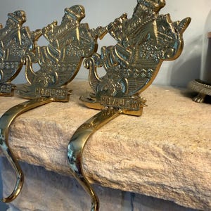 May include: A set of three polished brass stocking holders, each featuring a detailed relief of Santa Claus riding in a sleigh. The base of each sleigh is embossed with the text HO HO HO. Each holder includes a curved hook for hanging items.