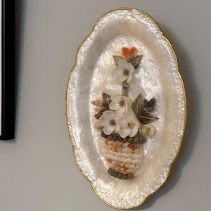 Peut inclure: Décoration murale ovale avec une bordure dorée. Elle présente un vase en coquillage rempli de fleurs blanches, de feuilles vertes et d'un petit cœur orange. Le fond est nacré, créant un effet scintillant.