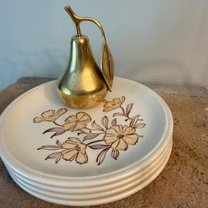 May include: A stack of three off-white ceramic plates featuring a brown floral pattern, topped with a polished brass pear-shaped decorative object that includes a curved stem and a single leaf extending from the top of the fruit.