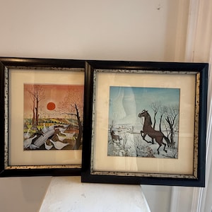 May include: Two square framed prints with black frames and silver inner borders. One print depicts a red sun over a field with white geese, while the other shows a brown horse rearing in a winter landscape with bare trees and a light blue sky.