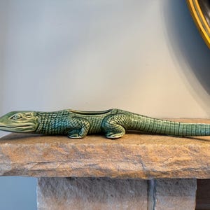 Peut inclure: Une jardinière en céramique verte en forme d'alligator. L'objet présente une surface texturée et écailleuse, une tête détaillée avec des yeux définis et une longue queue. Le dos de l'alligator est ouvert, créant un espace creux pour planter.