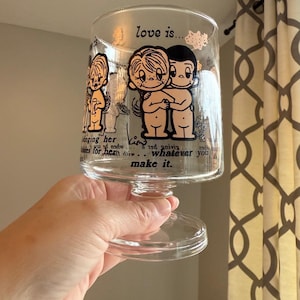 May include: Clear glass goblet with a wide bowl and a pedestal base. The glass is decorated with cartoon figures and the words "love is..." and "make it."