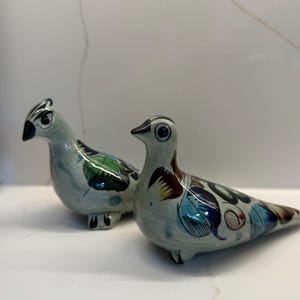 May include: Two ceramic bird figurines featuring a light grey base colour with hand-painted decorative patterns in shades of blue, green, brown, and black. Each bird has a smooth, glossy finish, a pointed beak, and detailed wing markings.