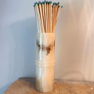 May include: A cylindrical stone holder with a polished cream and tan banded finish, featuring a raised horizontal ring detail around the centre. The holder is filled with a bundle of long wooden matches that have light blue tips.