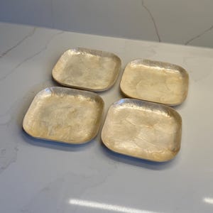 May include: A set of four square plates with rounded corners, featuring a shimmering, iridescent off-white finish with a layered, translucent shell-like texture across the surface of each individual plate, arranged in a two-by-two grid formation.