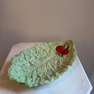 May include: A ceramic serving dish shaped like a textured green lettuce leaf. The dish features a glossy finish with a prominent central vein and ruffled edges. A small, bright red tomato with green leaves is attached to the right side of the leaf.