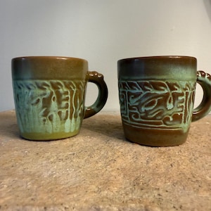 May include: Two ceramic mugs featuring a two-tone brown and light green glaze. Each mug has a cylindrical body with a thick, rounded handle. The exterior surface displays a textured, embossed pattern that wraps around the middle of the mugs.