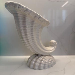 May include: A white ceramic cornucopia vase featuring a textured, shell-like surface pattern. The vessel has a wide, flared opening with a scalloped rim and rests on a rounded, ridged base that resembles a large seashell, creating a curved silhouette.