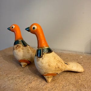 Peut inclure: Deux figurines d'oiseaux en céramique avec des têtes orange vif, des colliers bleu sarcelle foncé et des corps texturés blanc cassé. Chaque oiseau possède un petit bec sombre, des yeux ronds et une finition lisse sur la tête et le cou.
