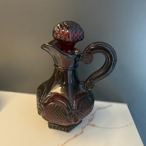 May include: A deep red glass decanter featuring a textured diamond pattern on its body and a matching stopper. The vessel has a curved handle with circular detailing and a flared spout, showcasing a rich, translucent ruby colour throughout the glass.