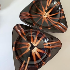 May include: Two triangular ashtrays with a glossy finish. The ashtrays are dark brown with a radial design in shades of orange and cream. Each ashtray has three built-in cigarette rests. The ashtrays are likely made of ceramic or glass.