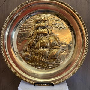 Peut inclure: Une assiette murale circulaire en laiton présentant un motif en relief d'un voilier à plusieurs mâts sur l'océan. L'assiette possède une finition métallique polie et une bordure décorative en forme de corde sur le pourtour extérieur.