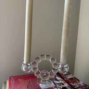 Puede incluir: Un candelabro de vidrio transparente con dos velas color crema. El soporte presenta un diseño circular de pequeñas esferas de vidrio y una base redonda. Las velas son altas y delgadas, agregando un toque de elegancia a la decoración.