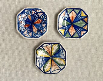 Puede incluir: Tres pequeños platos octogonales de cerámica con diseños pintados a mano. Cada plato presenta un patrón único en azul, naranja, amarillo y verde, con un borde blanco. Los platos están dispuestos sobre una tela de color neutro.