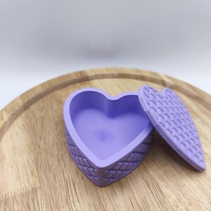 Peut inclure: Un récipient en forme de cœur violet clair avec un motif en losange texturé sur les côtés extérieurs et un couvercle assorti en forme de cœur présentant un design gaufré en grille, montré avec le couvercle retiré à côté du récipient ouvert.