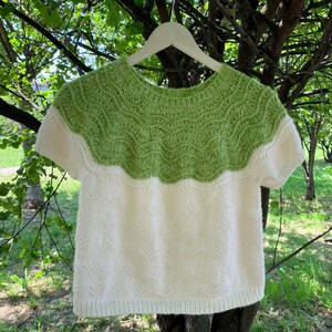 May include: A short-sleeved knit sweater featuring a round neckline and a two-tone color scheme. The upper yoke displays a vibrant lime green hue with a textured wave pattern, while the lower body is a solid off-white with a subtle diamond stitch.