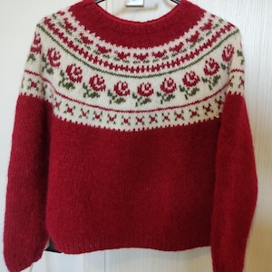 May include: A red sweater with a white and green floral pattern across the chest and upper sleeves. The sweater has a crew neck and long sleeves. The floral design includes red roses and green leaves. The sweater is hanging on a hanger.
