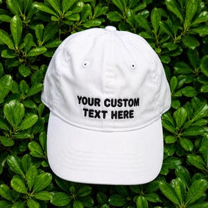 Peut inclure: Une casquette de baseball blanche avec le texte noir brodé "YOUR CUSTOM TEXT HERE" centré sur le panneau avant. La casquette présente une calotte structurée, une visière incurvée et un bouton sur le dessus, offrant un design épuré.