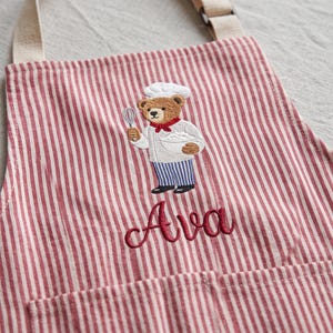 May include: A red and white striped apron featuring an embroidered brown teddy bear dressed as a chef, holding a whisk and a bowl. The name Ava is embroidered in red cursive script below the bear. The apron has beige adjustable neck straps.
