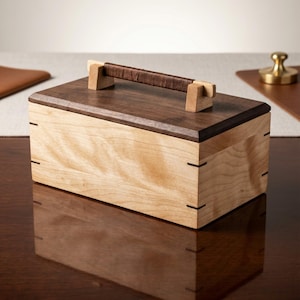 May include: A rectangular wooden box featuring a light-colored body with visible grain and a dark brown lid. The lid includes a central handle wrapped in dark brown material, secured by two light-colored wooden supports on either side.