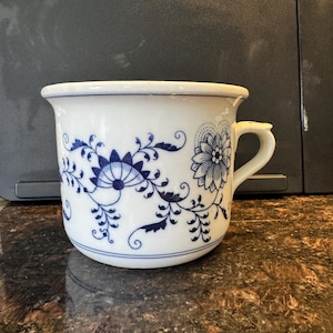 Op de afbeelding: Witte keramische mok met een blauw bloemmotief. De mok heeft een handvat en een gedetailleerd ontwerp van bloemen en bladeren. Het patroon herhaalt zich rond de buitenkant van de mok. Voor dranken.