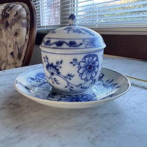 Op de afbeelding: Een wit en blauw porseleinen suikerpotje met deksel, rustend op een bijpassende schotel. De kom en schotel hebben een bloemmotief in blauw. De set staat op een marmeren oppervlak, met een stoel en een raam op de achtergrond.