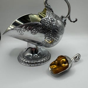Puede incluir: Un azucarero plateado con una cuchara a juego. El azucarero tiene un pico curvo, detalles florales ornamentados y un asa decorativa. La cuchara tiene un asa pequeña y un interior dorado. El conjunto está sobre una superficie blanca.
