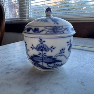 Puede incluir: Un tarro de cerámica blanco con tapa, adornado con patrones florales azules. El tarro tiene forma redondeada y un pequeño pomo en la parte superior de la tapa. El diseño azul y blanco es un estilo clásico, adecuado para guardar objetos pequeños.