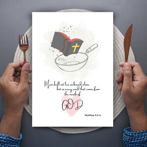 Könnte beinhalten: Ein weißer Druck mit einer Bibel, einer Bratpfanne und dem Text "Der Mensch lebt nicht vom Brot allein, sondern von jedem Wort, das aus dem Mund Gottes kommt." Auf beiden Seiten befinden sich eine Gabel und ein Messer.