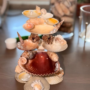 May include: A tiered decorative stand constructed from various seashells and small white pearls. The structure features multiple levels of stacked scallop, conch, and spiral shells in shades of cream, tan, and deep reddish-brown, arranged in a circular formation.