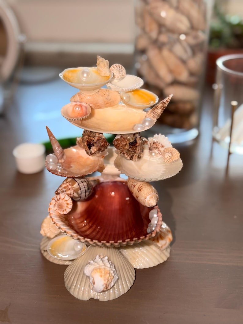 May include: A tiered decorative stand constructed from various seashells and small white pearls. The structure features multiple levels of stacked scallop, conch, and spiral shells in shades of cream, tan, and deep reddish-brown, arranged in a circular formation.