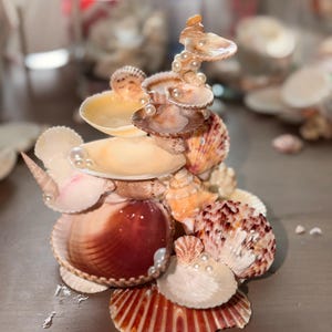 May include: A decorative sculpture constructed from various natural seashells in shades of brown, tan, cream, and white. The structure features multiple stacked shells arranged in a tiered formation, accented with small, round, white faux pearls.