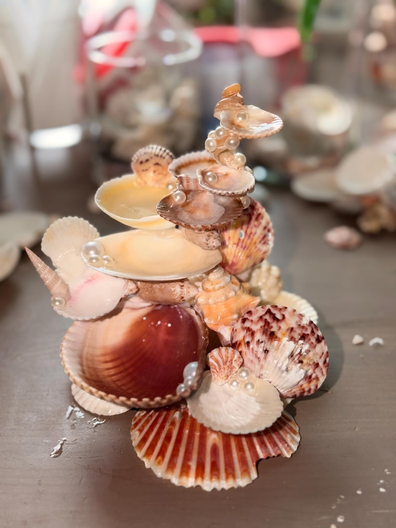 May include: A decorative sculpture constructed from various natural seashells in shades of brown, tan, cream, and white. The structure features multiple stacked shells arranged in a tiered formation, accented with small, round, white faux pearls.