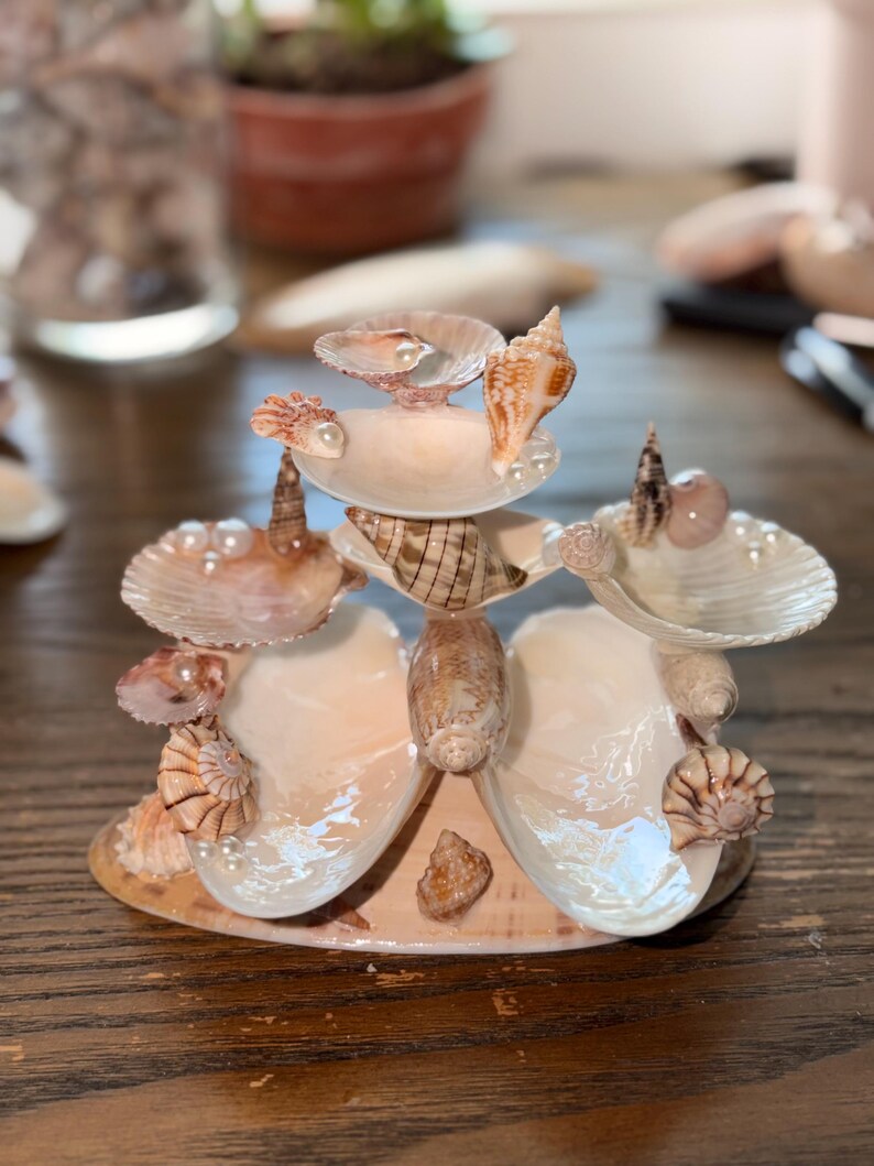 May include: A tiered decorative display stand constructed from various seashells and small white pearls. The structure features multiple shell platforms arranged in a vertical formation, with spiraled and bivalve shells serving as supports.
