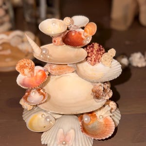 May include: A tiered decorative stand constructed from various seashells, including white, tan, and orange scallop and conch shells. The structure features multiple levels adorned with small white faux pearls tucked into the shell openings.