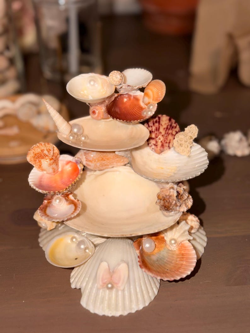 May include: A tiered decorative stand constructed from various seashells, including white, tan, and orange scallop and conch shells. The structure features multiple levels adorned with small white faux pearls tucked into the shell openings.