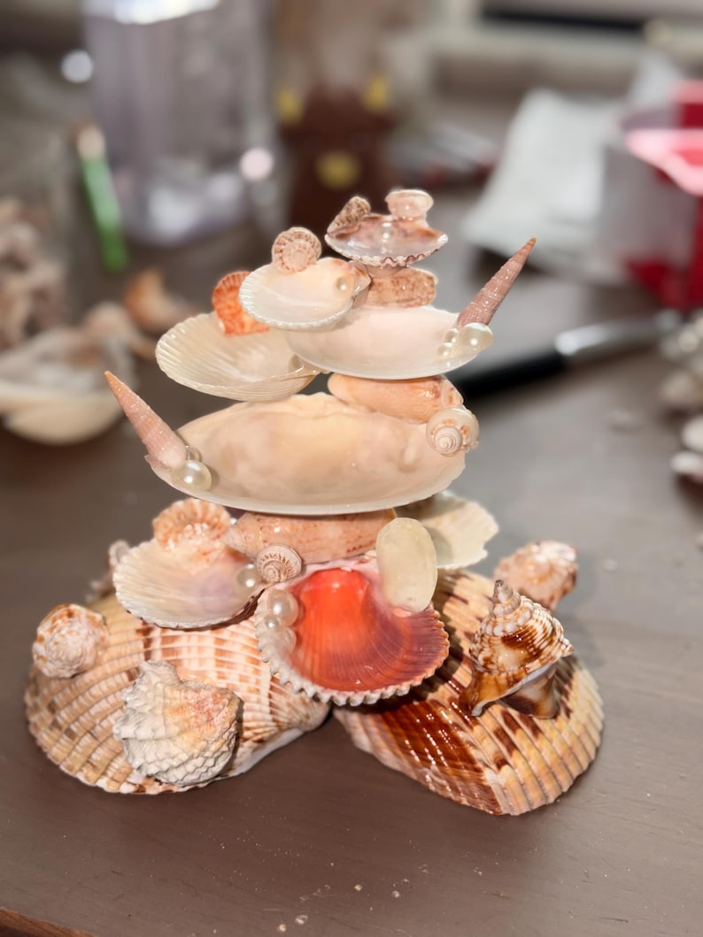 May include: A decorative sculpture constructed from various seashells, including large brown striped bivalves at the base, smaller white fan-shaped shells, spiral shells, and small white pearls arranged in a tiered, stacked formation.