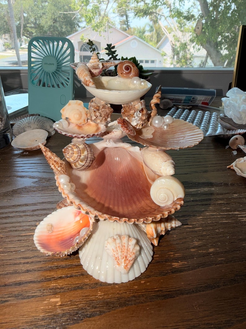 May include: A tiered decorative sculpture constructed from various seashells and white pearls. The structure features multiple levels of interconnected bivalve and spiral shells in shades of cream, tan, and brown, arranged to form a central display piece.
