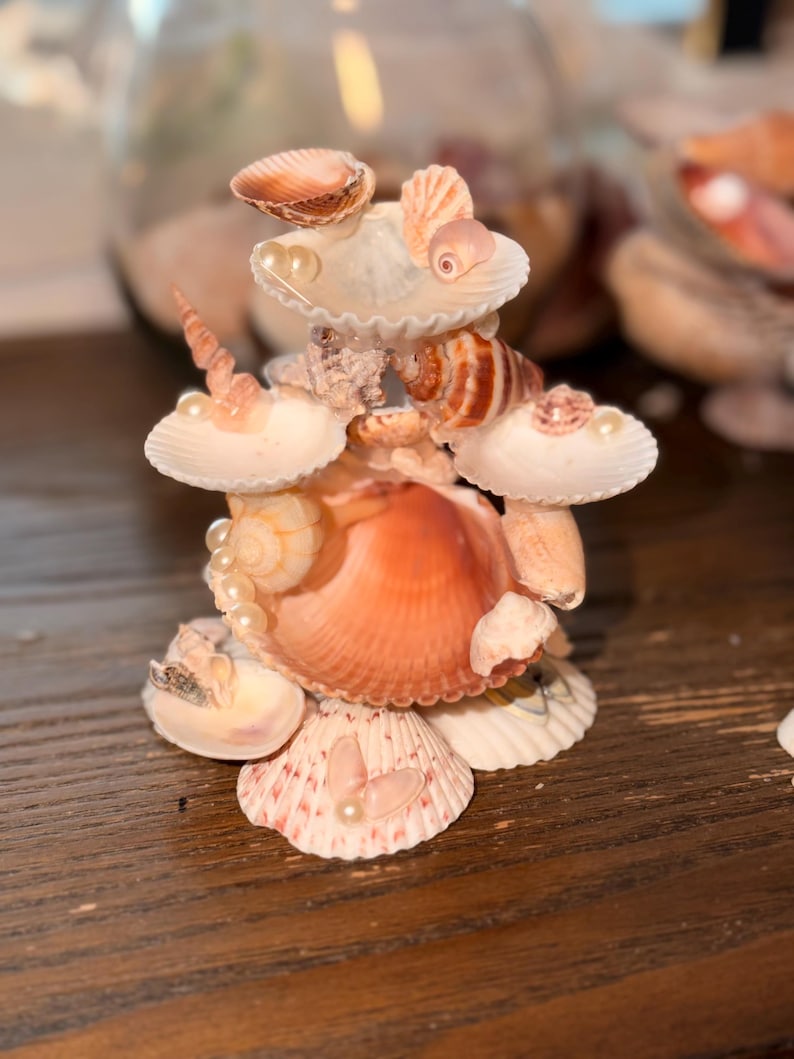 May include: A decorative sculpture constructed from various seashells and small white faux pearls. The structure features multiple tiers of stacked scallop and spiral shells in shades of cream, tan, and soft orange, arranged in a vertical formation.