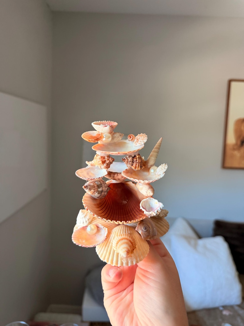 May include: A decorative sculpture constructed from various natural seashells in shades of tan, cream, and brown, arranged in a tiered, vertical formation. The structure incorporates small white spherical pearls nestled within the shell openings.