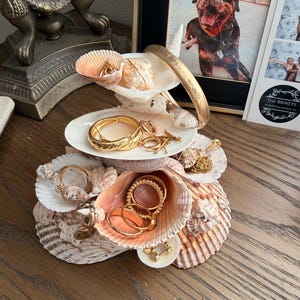 Può includere: Un portagioielli a conchiglia a più livelli che mostra vari anelli, bracciali e collane d'oro. Il supporto è realizzato con conchiglie naturali e poggia su una superficie di legno. Una foto incorniciata e una lampada decorativa sono sullo sfondo.